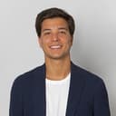 A close-up portrait of a young man with short dark brown hair and a warm, friendly smile, wearing a white crew-neck t-shirt under a navy blue textured blazer, set against a plain light gray studio background with soft, even lighting that highlights his facial features and creates a professional, approachable look.